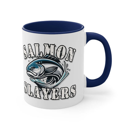 A white custom Salmon Slayers - Fierce Flash - Accent Coffee Mug, 11oz with a blue interior and C-handle features the design "Salmon Slayers" with a stylized illustration of a salmon in the center. The mug, showcasing an eye-catching color contrast, is placed on a wooden surface next to a small macaron, coffee beans, and a spoon.