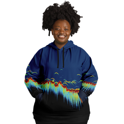 A person wearing a Sonar Scan Plus-size Hoodie, featuring vibrant abstract wave patterns, stands smiling against a black background. Their curly hair complements the playful design, exuding a cheerful demeanor that makes this piece an ideal addition to any fishing wardrobe.