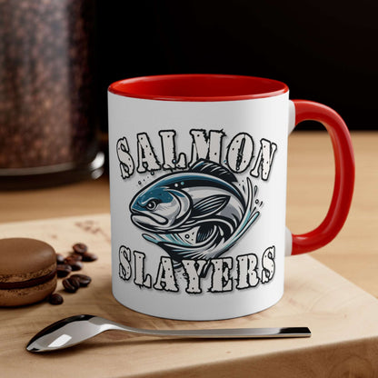 A white custom Salmon Slayers - Fierce Flash - Accent Coffee Mug, 11oz with a blue interior and C-handle features the design "Salmon Slayers" with a stylized illustration of a salmon in the center. The mug, showcasing an eye-catching color contrast, is placed on a wooden surface next to a small macaron, coffee beans, and a spoon.