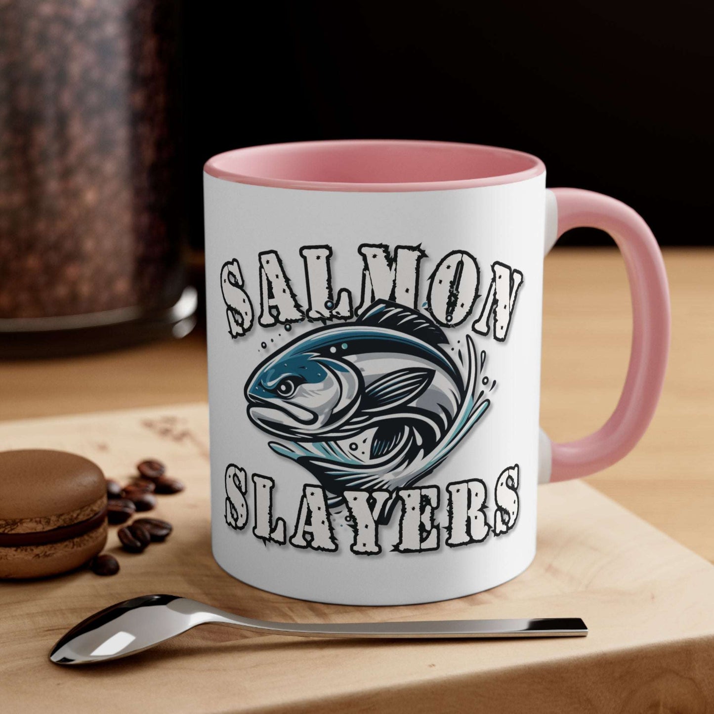 A white custom Salmon Slayers - Fierce Flash - Accent Coffee Mug, 11oz with a blue interior and C-handle features the design "Salmon Slayers" with a stylized illustration of a salmon in the center. The mug, showcasing an eye-catching color contrast, is placed on a wooden surface next to a small macaron, coffee beans, and a spoon.