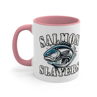 A white custom Salmon Slayers - Fierce Flash - Accent Coffee Mug, 11oz with a blue interior and C-handle features the design "Salmon Slayers" with a stylized illustration of a salmon in the center. The mug, showcasing an eye-catching color contrast, is placed on a wooden surface next to a small macaron, coffee beans, and a spoon.