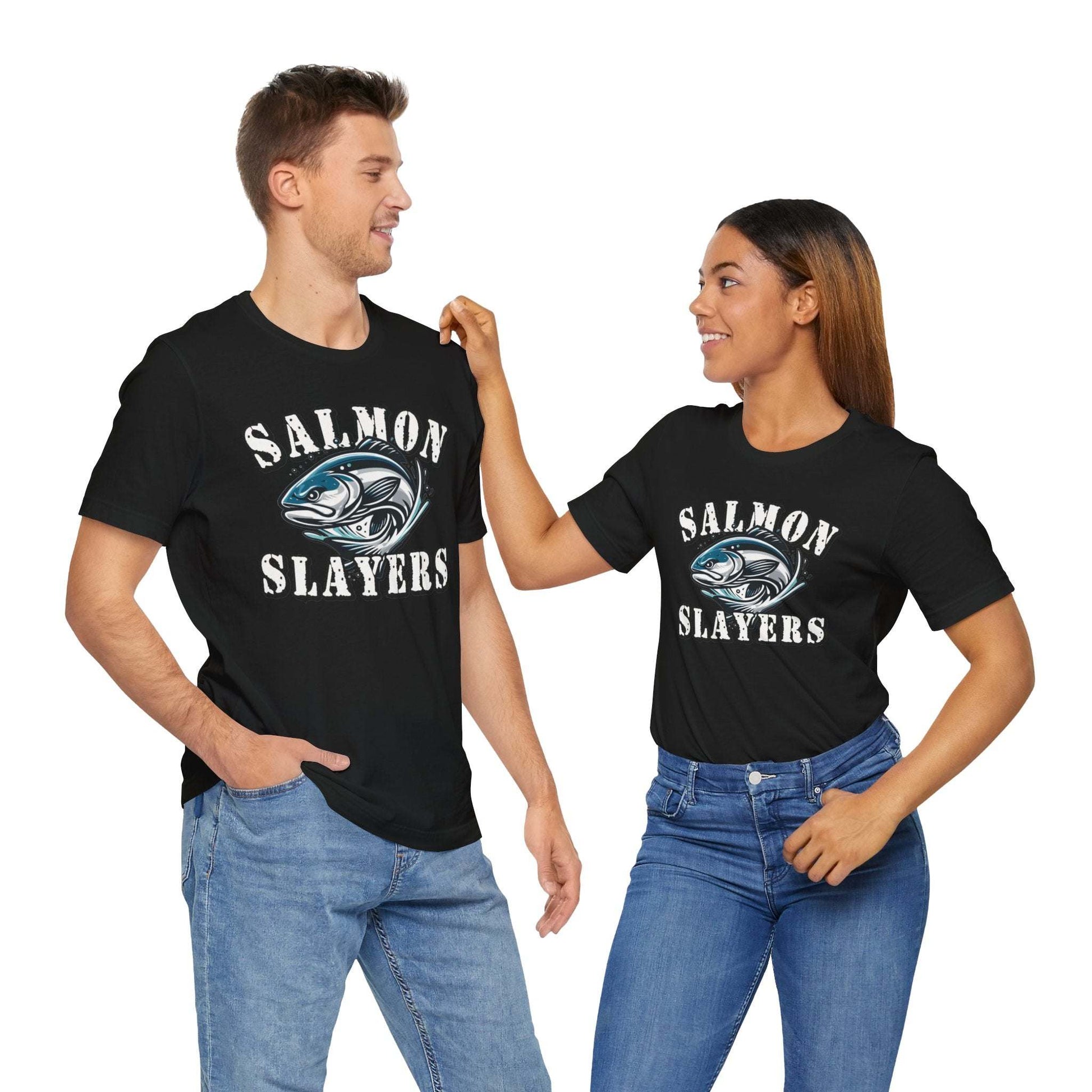 A blue unisex T-shirt made from 100% Airlume cotton featuring the text "Salmon Slayers" in bold, white, distressed letters. Below, an illustration of a fierce-looking fish with sharp teeth and dynamic lines suggests movement. Enjoy fast shipping on this striking design: **Slamon Slayers - Fierce Flash - T-Shirt**.