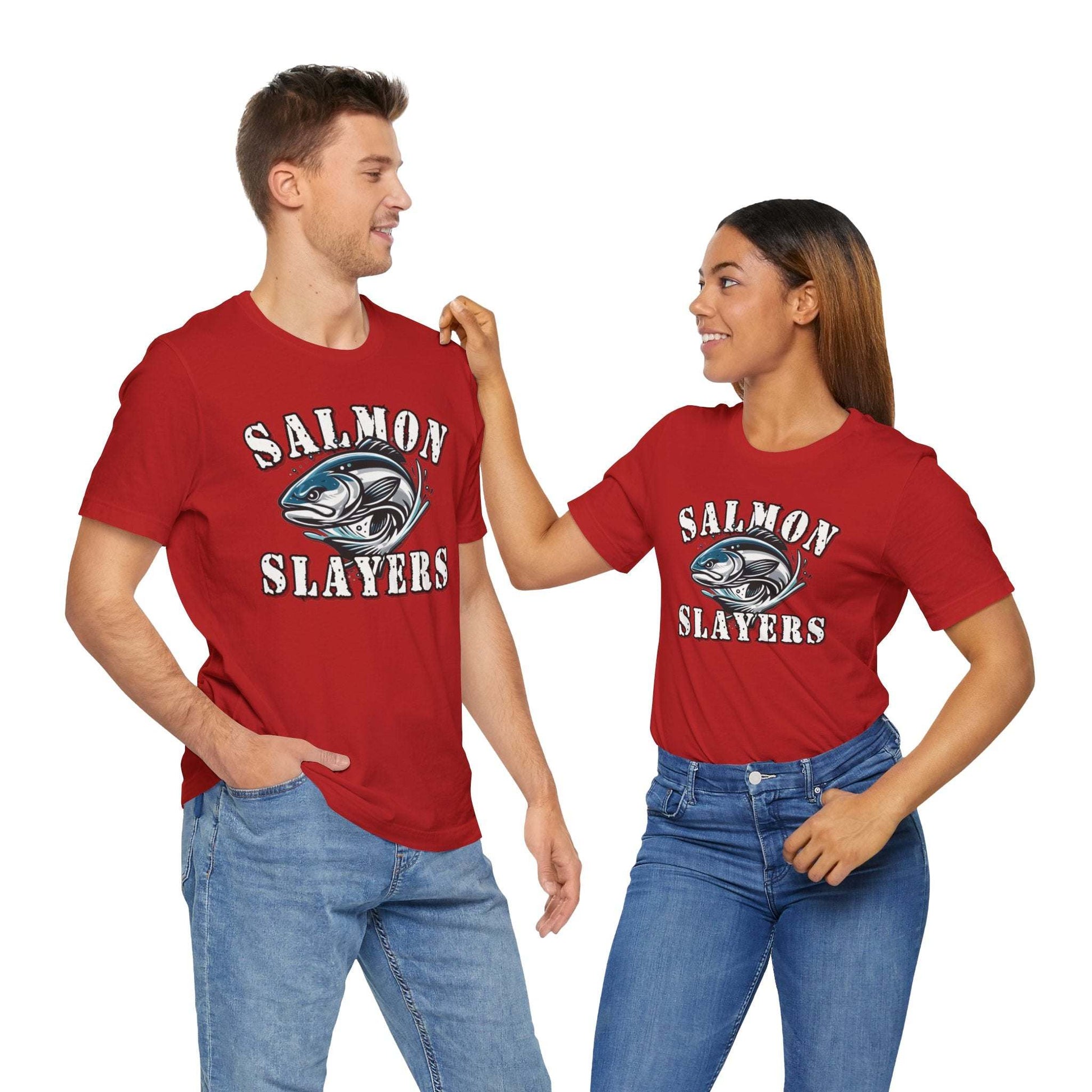 A blue unisex T-shirt made from 100% Airlume cotton featuring the text "Salmon Slayers" in bold, white, distressed letters. Below, an illustration of a fierce-looking fish with sharp teeth and dynamic lines suggests movement. Enjoy fast shipping on this striking design: **Slamon Slayers - Fierce Flash - T-Shirt**.