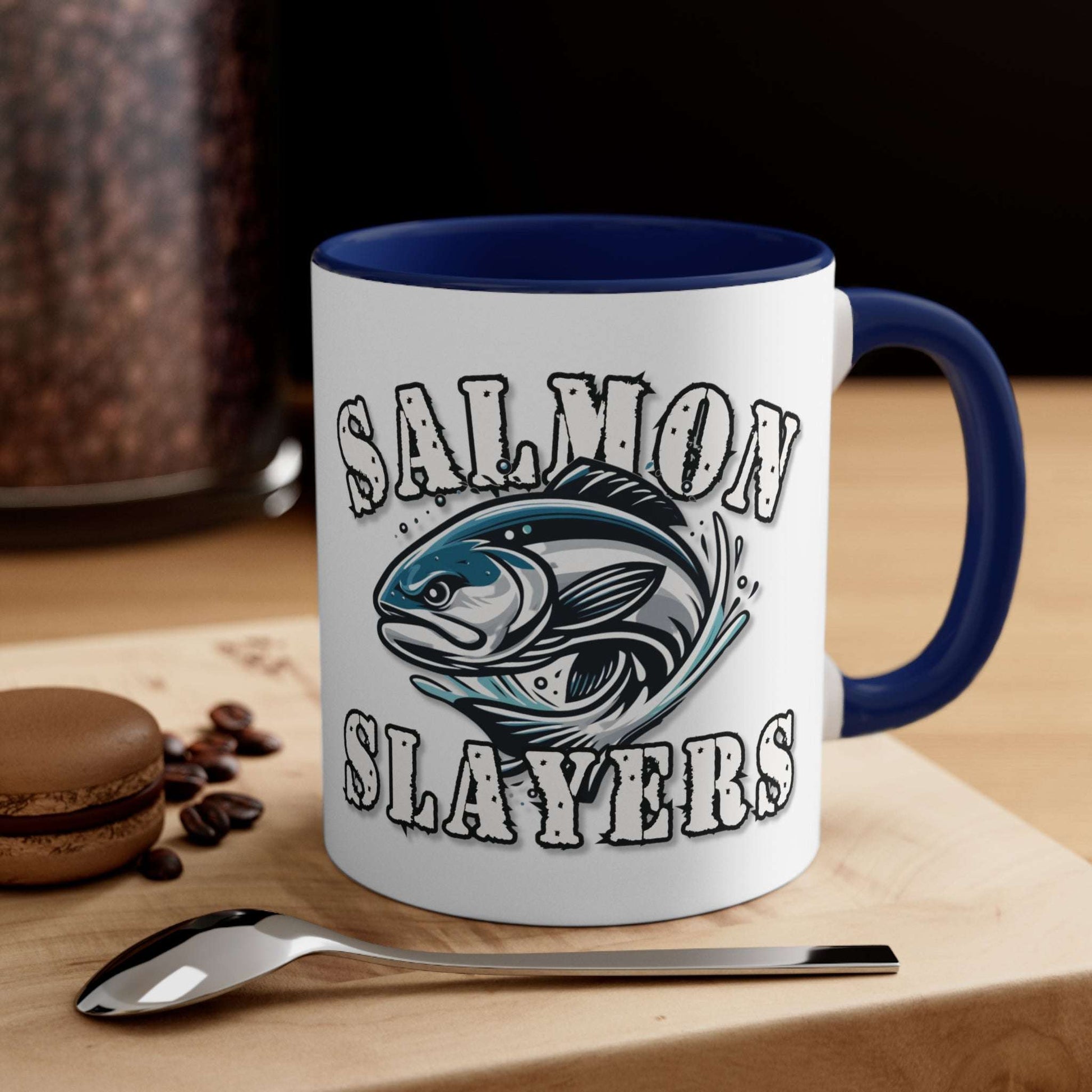 A white custom Salmon Slayers - Fierce Flash - Accent Coffee Mug, 11oz with a blue interior and C-handle features the design "Salmon Slayers" with a stylized illustration of a salmon in the center. The mug, showcasing an eye-catching color contrast, is placed on a wooden surface next to a small macaron, coffee beans, and a spoon.