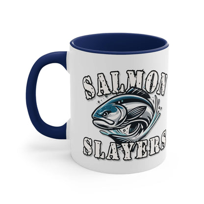 A white custom Salmon Slayers - Fierce Flash - Accent Coffee Mug, 11oz with a blue interior and C-handle features the design "Salmon Slayers" with a stylized illustration of a salmon in the center. The mug, showcasing an eye-catching color contrast, is placed on a wooden surface next to a small macaron, coffee beans, and a spoon.