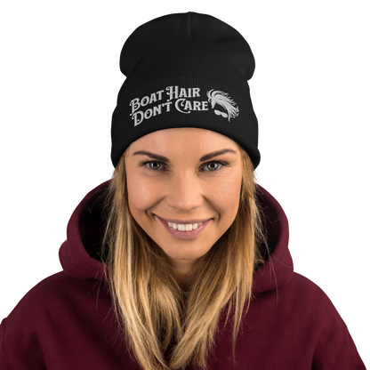 A cozy, dark green beanie with a folded brim showcases the embroidered white text "BEANIE - BOAT HAIR DON'T CARE" alongside a whimsical design of windblown hair and sunglasses.