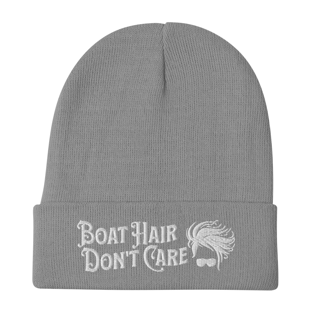 A cozy, dark green beanie with a folded brim showcases the embroidered white text "BEANIE - BOAT HAIR DON'T CARE" alongside a whimsical design of windblown hair and sunglasses.