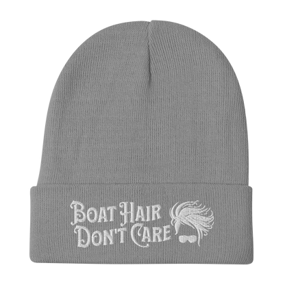 A cozy, dark green beanie with a folded brim showcases the embroidered white text "BEANIE - BOAT HAIR DON'T CARE" alongside a whimsical design of windblown hair and sunglasses.